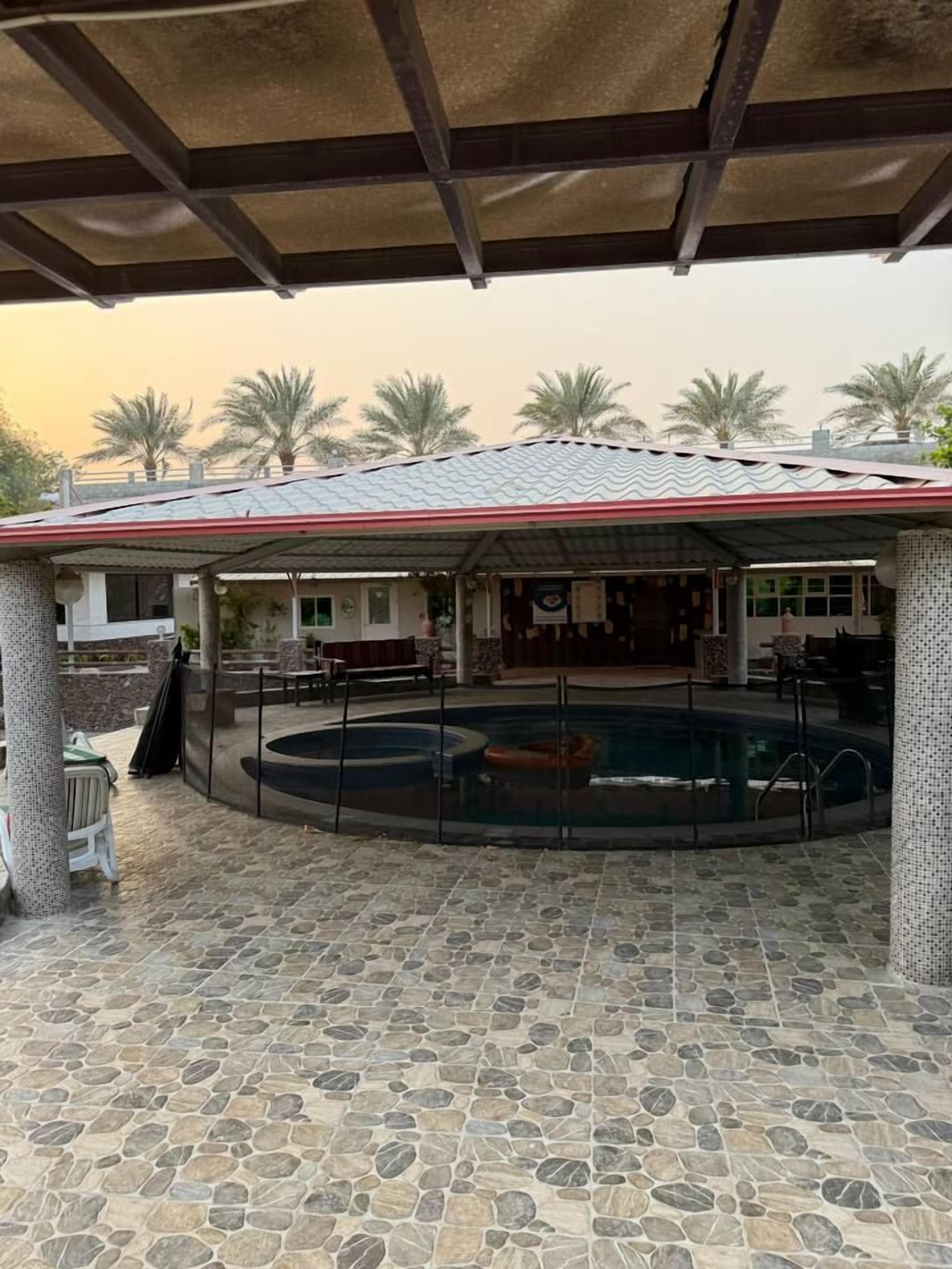 A covered outdoor area with a pool, stone flooring, and a building with a tiled roof in the background.