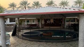 A covered outdoor area with a pool, stone flooring, and a building with a tiled roof in the background.