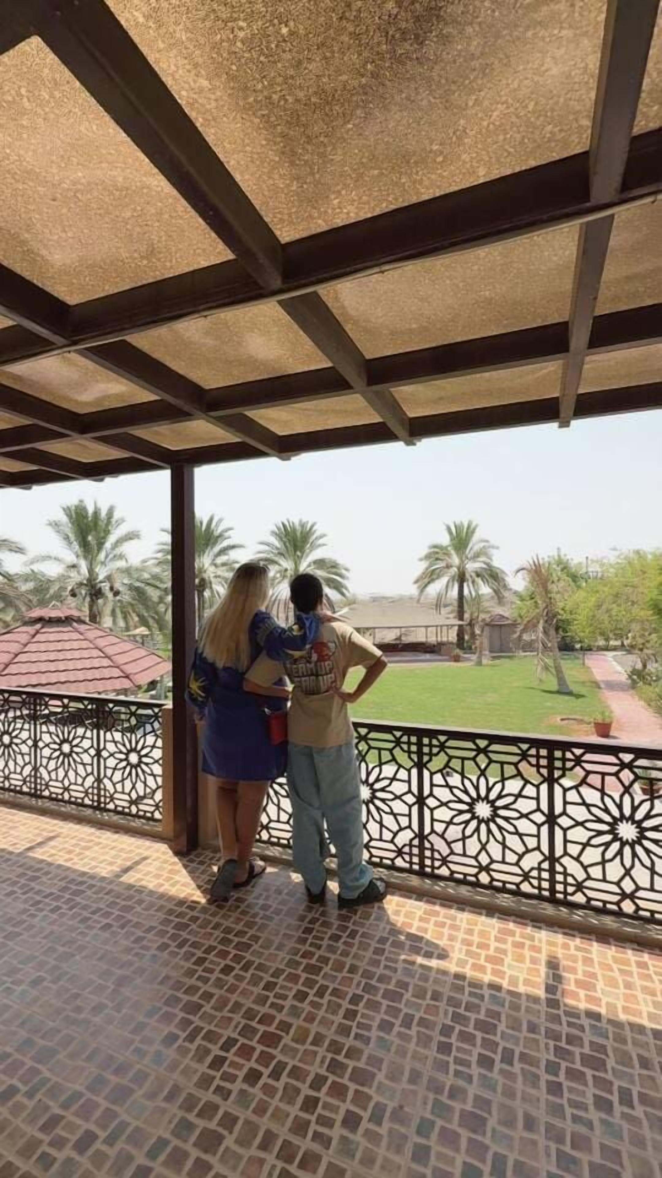 A covered outdoor area with a tiled floor, a metal railing, and a view of a green landscape with palm trees.