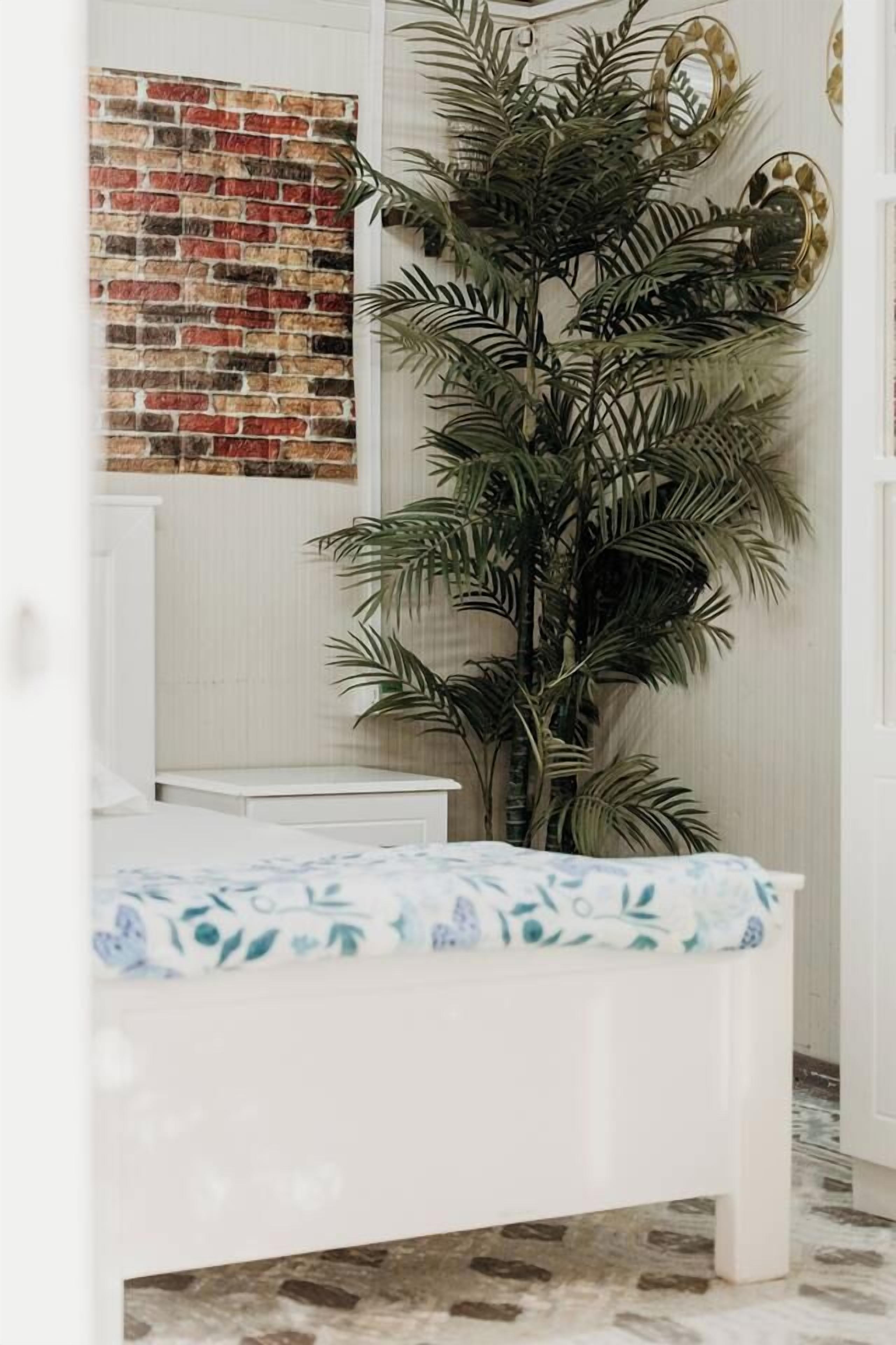 Una habitación con un banco blanco, una palmera artificial grande, un muro con diseño de ladrillo y un espejo.