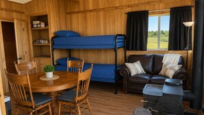 A cozy cabin interior with bunk beds, a dining table with chairs, a leather sofa, and a wood-burning stove.