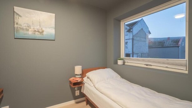 Um quarto com uma cama de solteiro, uma mesa de cabeceira com luminária, uma janela com vista para um edifício e um quadro na parede.