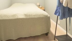 Quarto pequeno com cama, janela, cabideiro e uma mesinha.