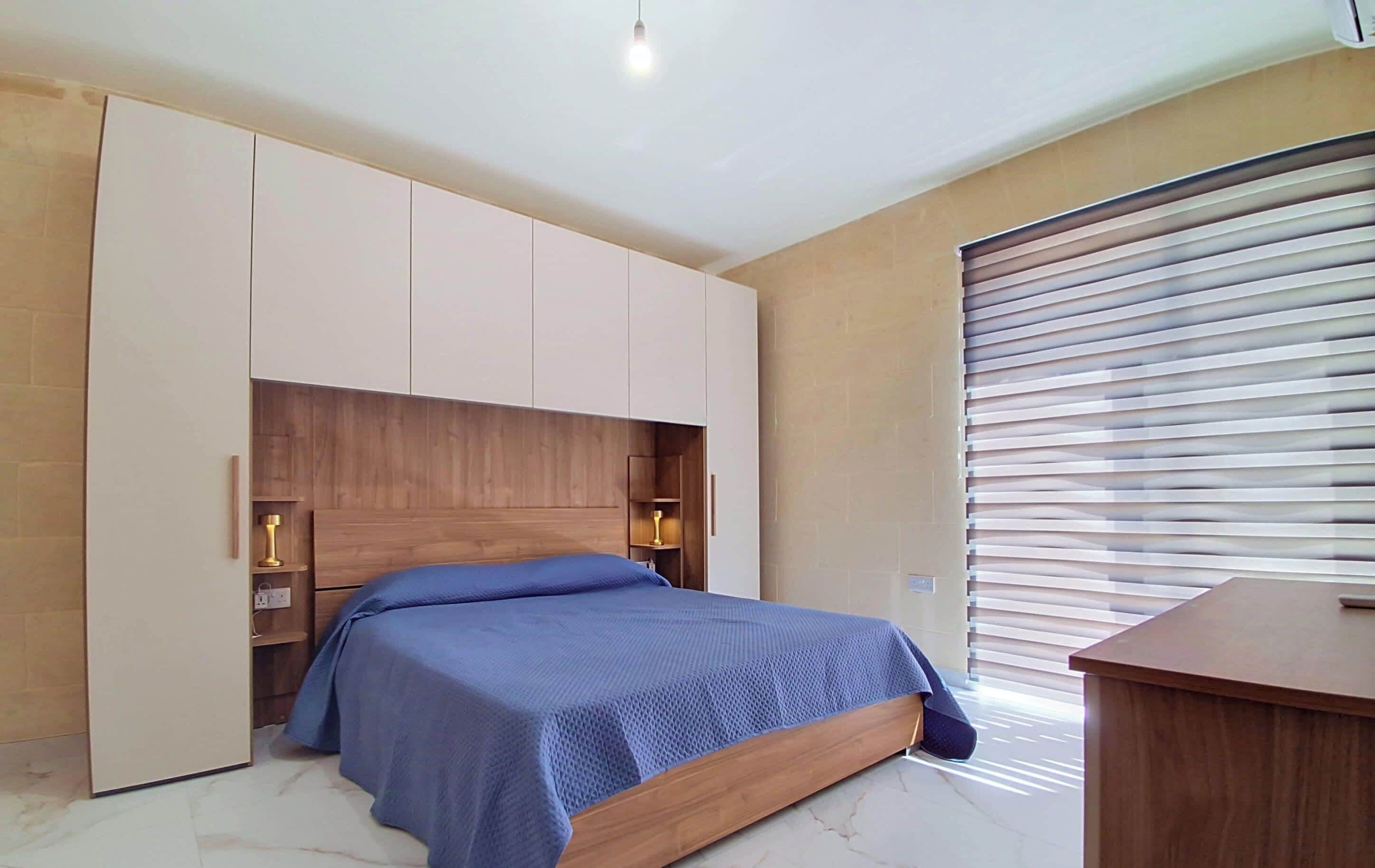 Une chambre à coucher comprenant un lit, une armoire intégrée, des lampes de chevet et une fenêtre avec des stores.