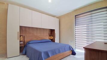 Ein Schlafzimmer mit Bett, integriertem Kleiderschrank, Nachttischlampen und einem Fenster mit Jalousien.