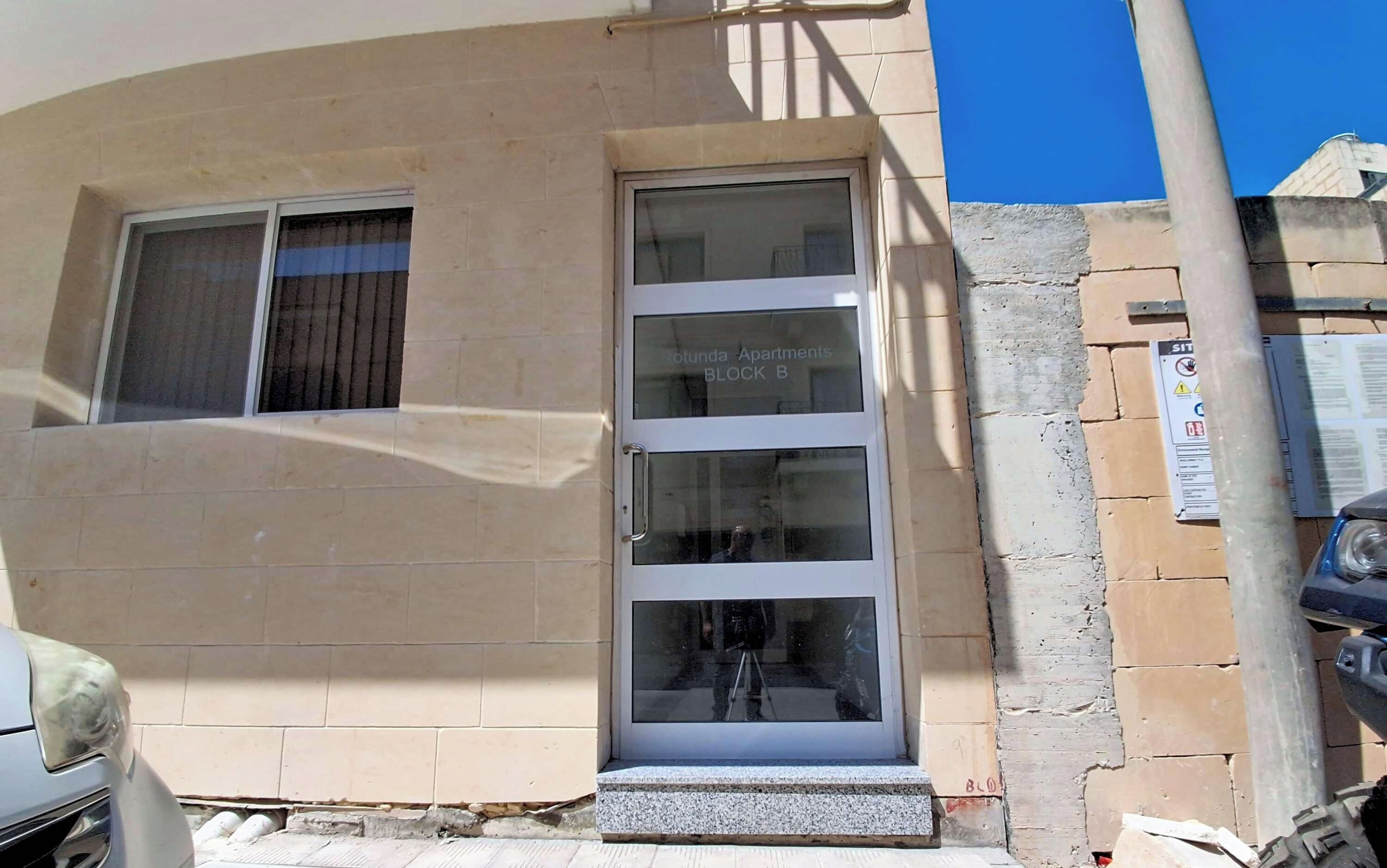 Fachada de un edificio con una ventana transparente, una puerta visible y un letrero que indica 'LOTUNDA APARTMENTS BLOCK B'.