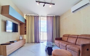 A modern living room with a flat-screen TV, a glass coffee table, a leather sofa, and a built-in TV shelf. - Spacious 2BR Family Apt in Central Gozo Location (Ix-Xewkija)
