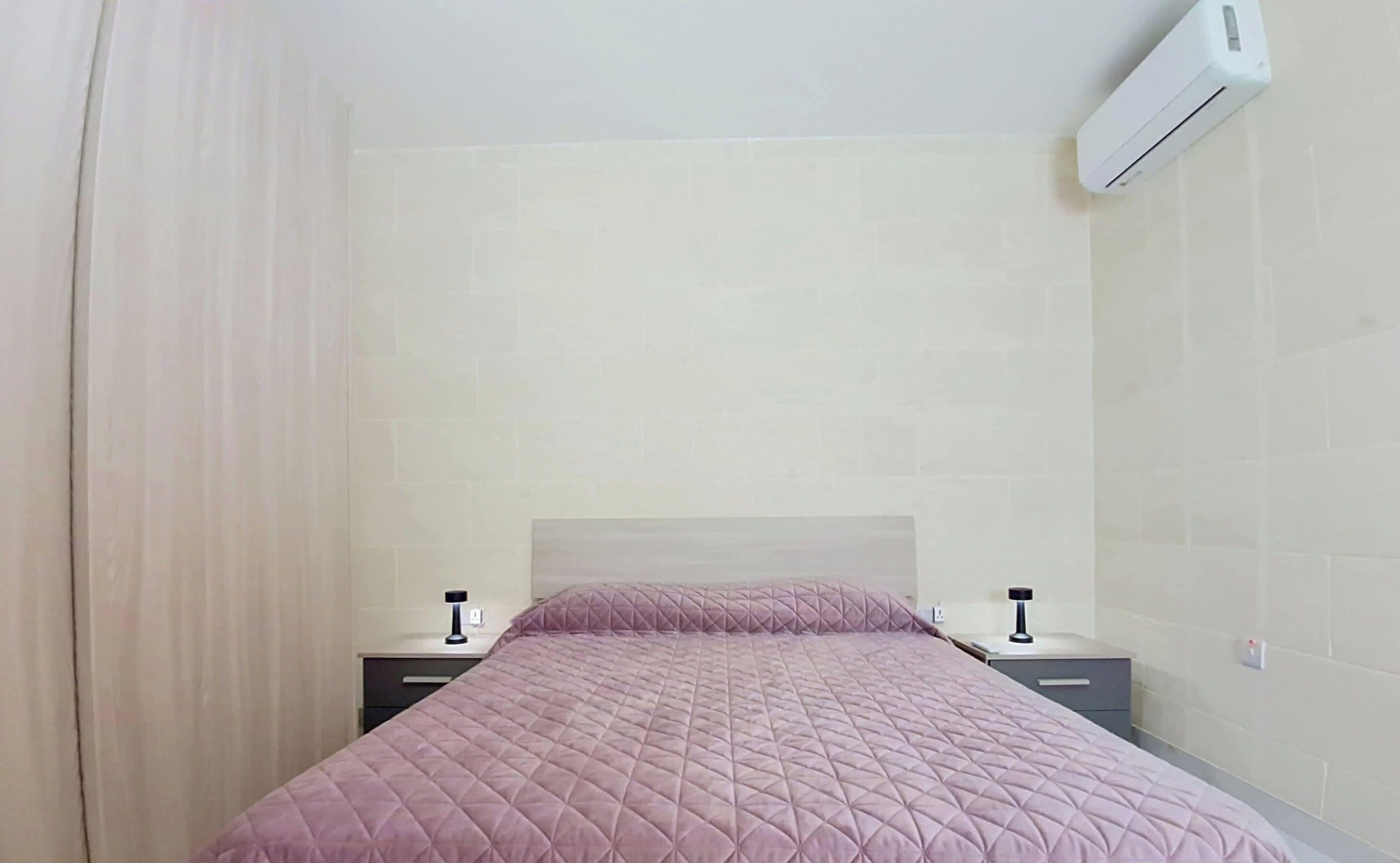 A bedroom with a bed, nightstands, an air conditioning unit, and a curtain.