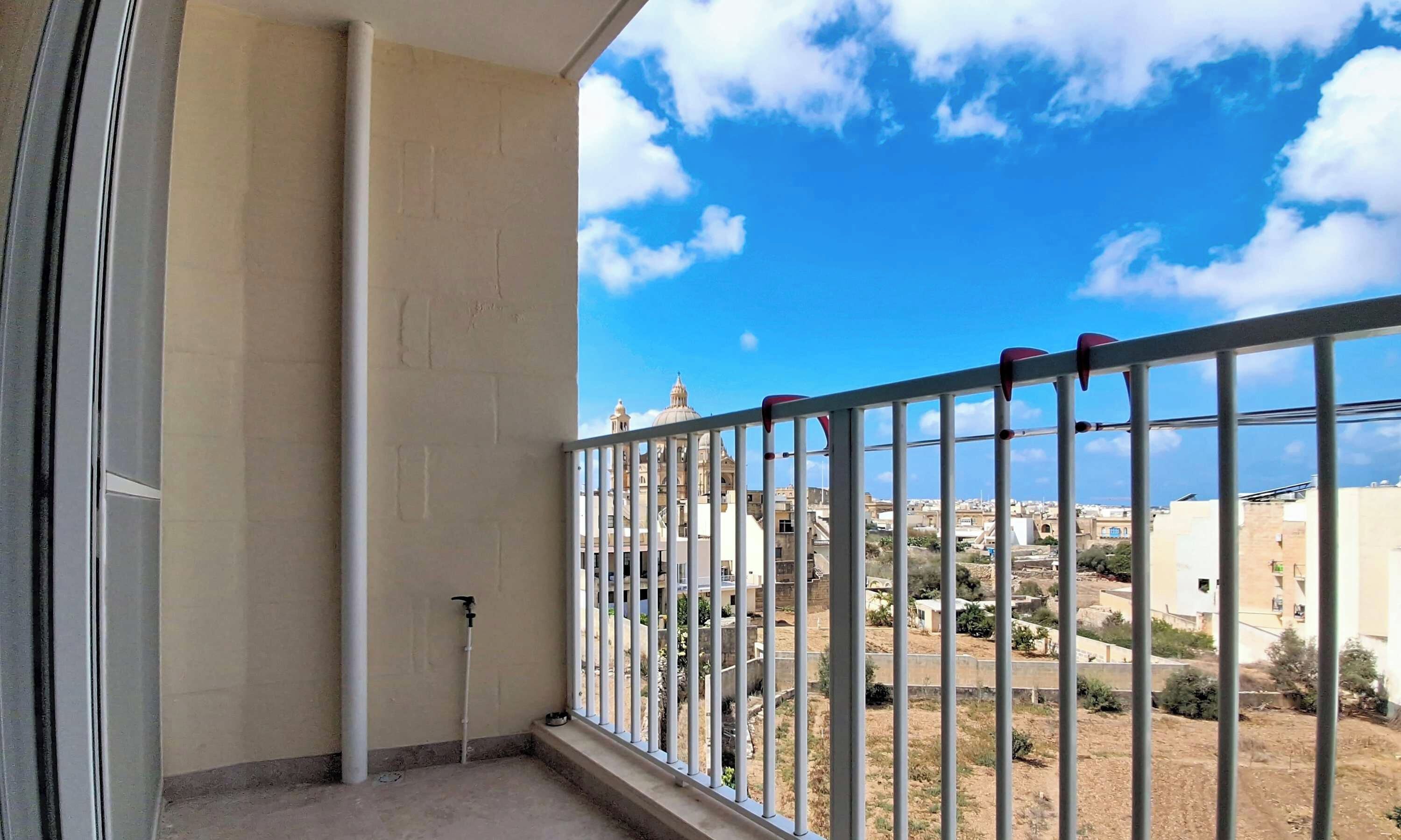 Un balcón con barandilla metálica, una tubería blanca y vistas a edificios y una estructura en forma de cúpula a lo lejos.