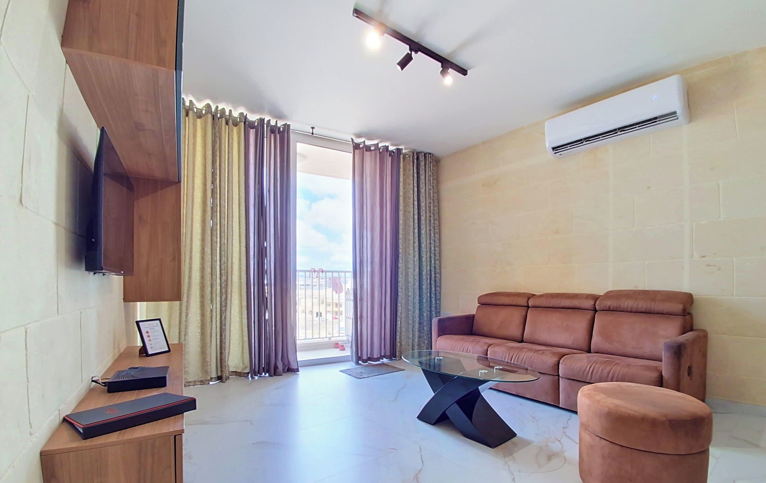 Una sala de estar con un sofá marrón, una mesa de centro de vidrio, un televisor montado en la pared y un equipo de aire acondicionado.