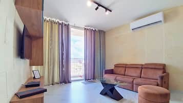 Una sala de estar con un sofá marrón, una mesa de centro de vidrio, un televisor montado en la pared y un equipo de aire acondicionado.