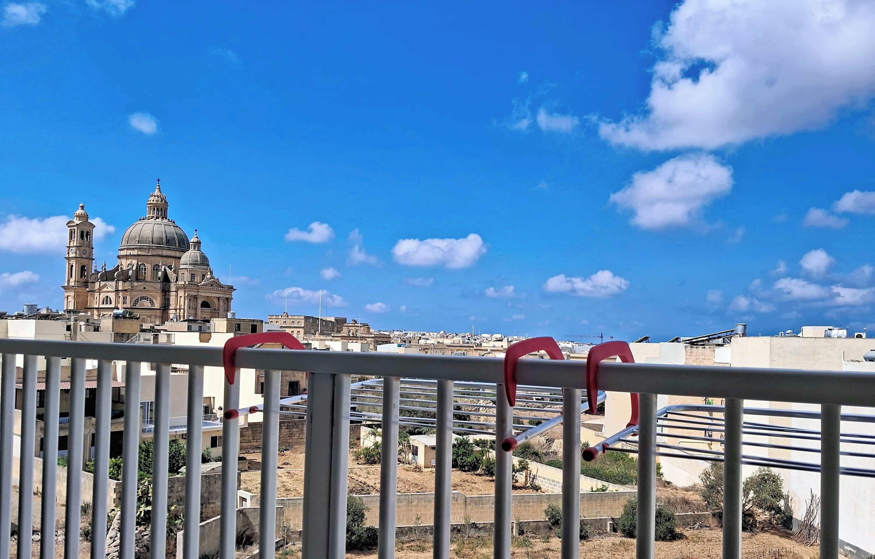 A view of a cityscape with a prominent church with a dome and bell tower, a clear blue sky, and a metal railing in the foreground.