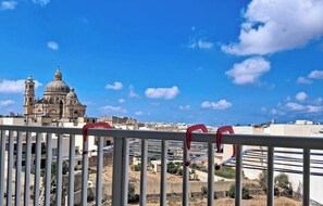 A view of a cityscape with a prominent church with a dome and bell tower, a clear blue sky, and a metal railing in the foreground. - Spacious 2BR Family Apt in Central Gozo Location (Ix-Xewkija)