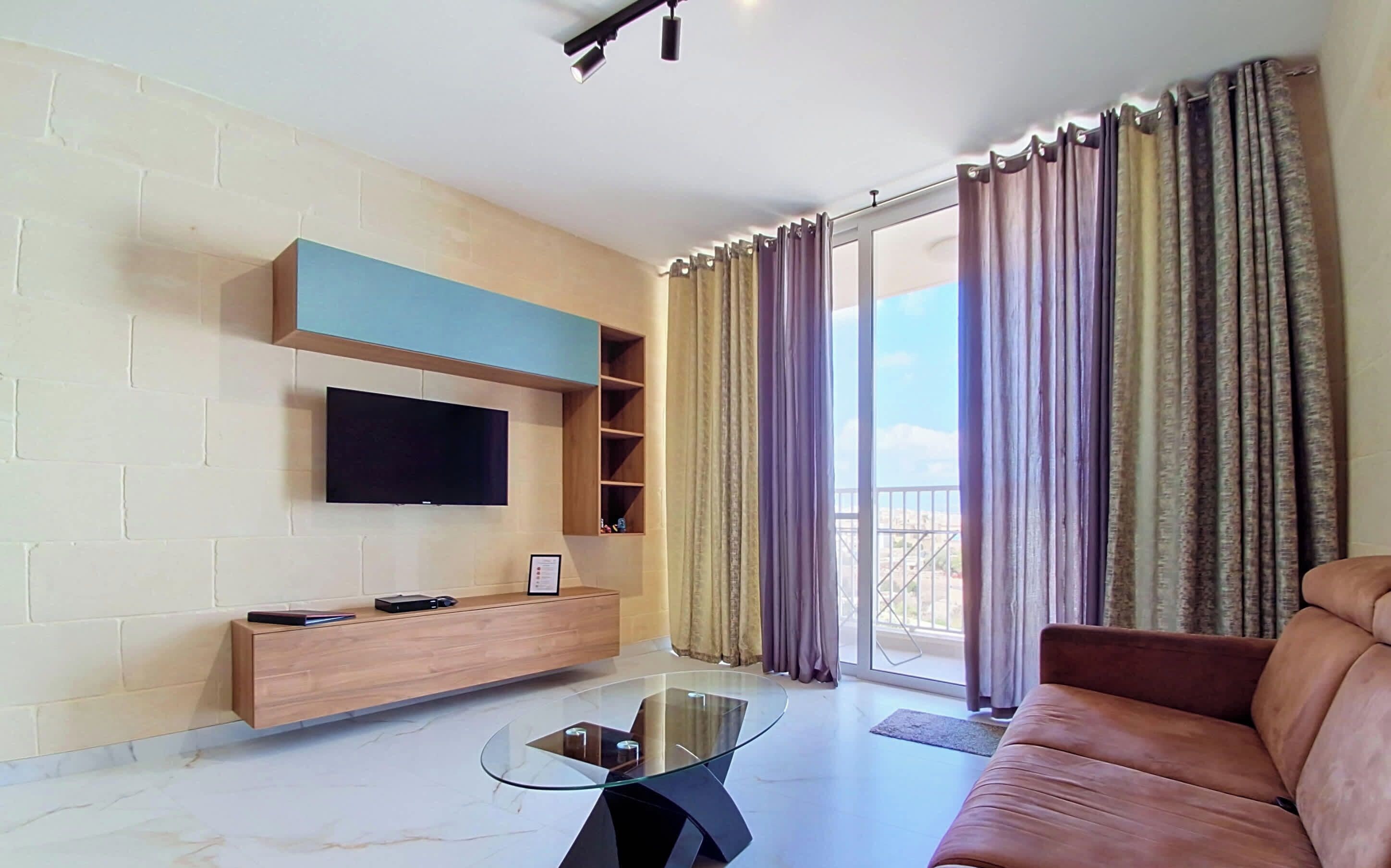 Una sala de estar moderna con televisor de pantalla plana, una mesa de centro de vidrio, un sofá y un balcón con cortinas.
