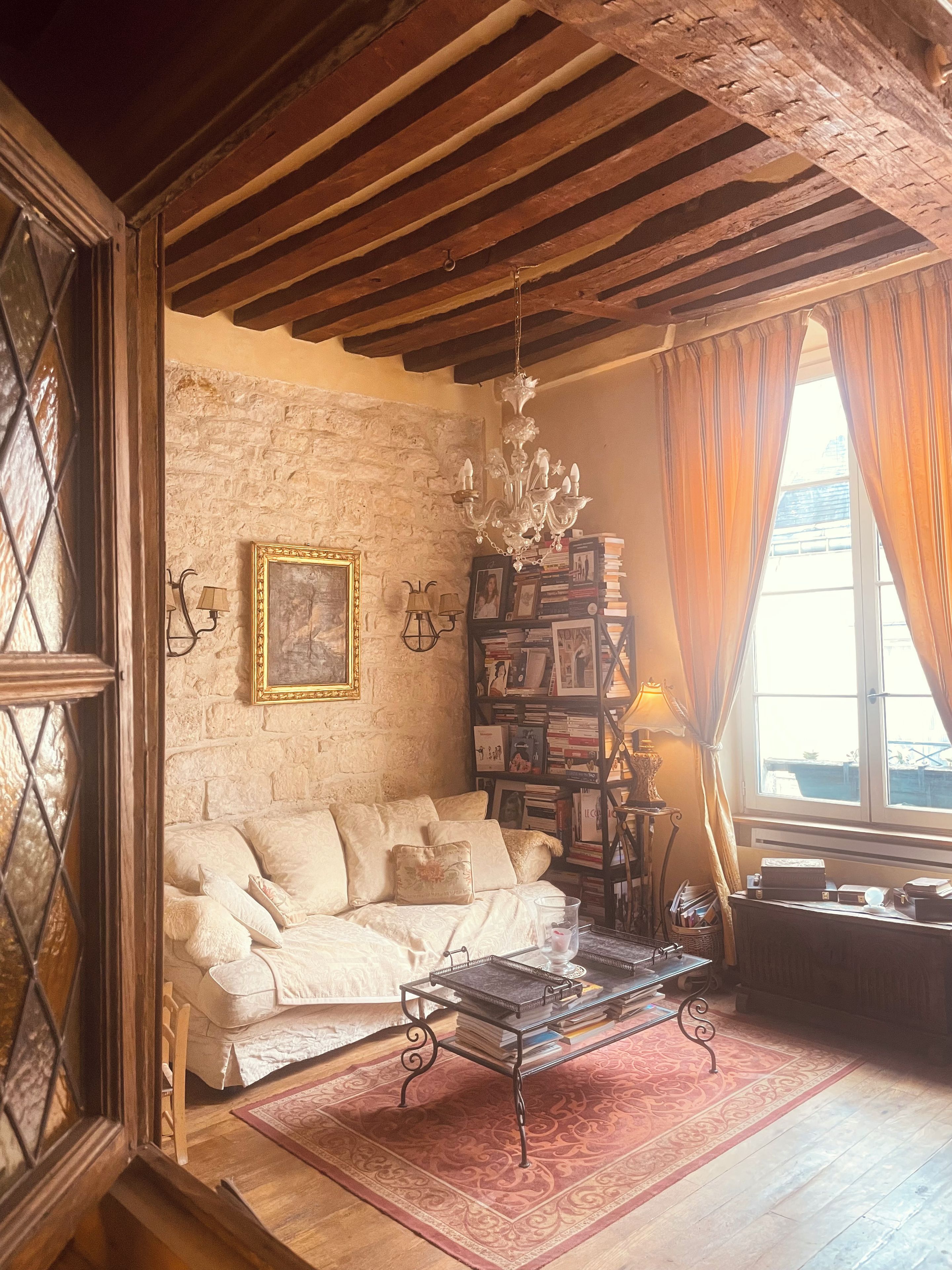 A cozy living room with a white sofa, a bookshelf, a chandelier, and a large window with curtains.