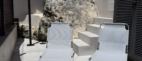 Dos sillones de descanso con estructura metálica y respaldo de malla, ubicados en un patio de concreto junto a un muro de piedra y un edificio con fachada color crema.