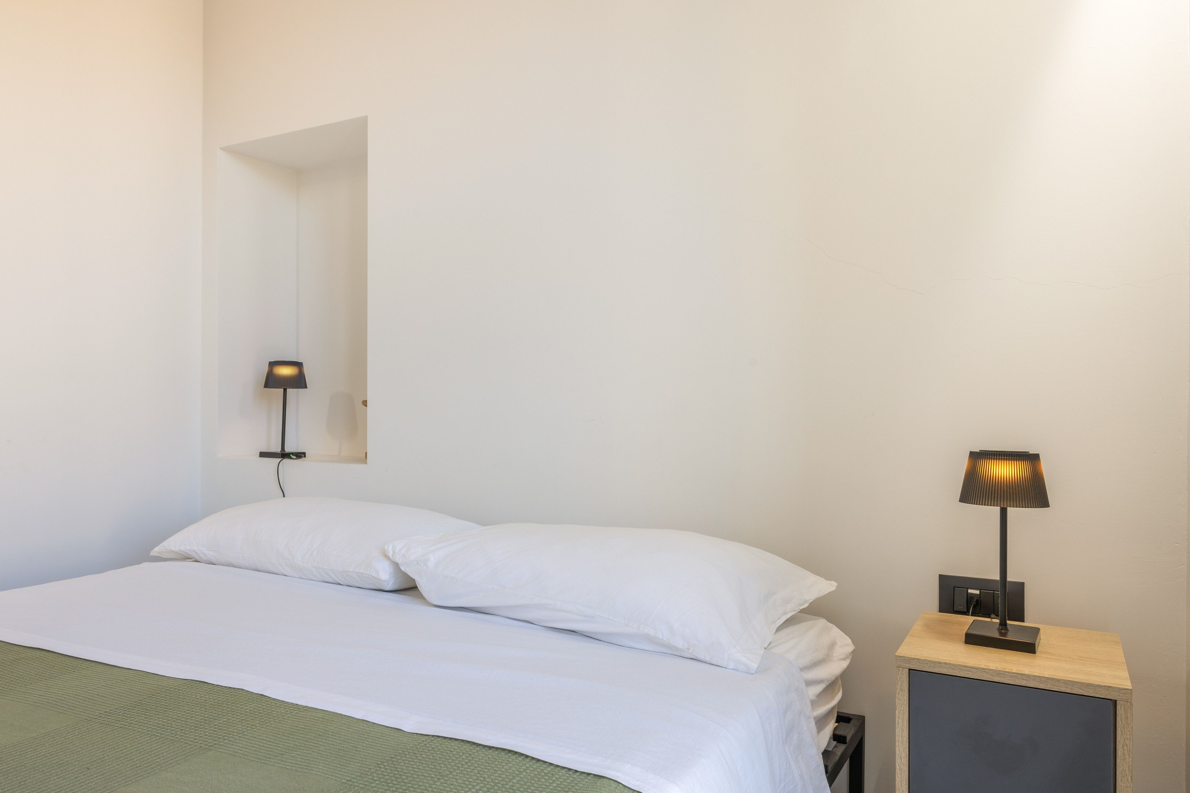 Quarto simples com uma cama, mesa de cabeceira e luminária.