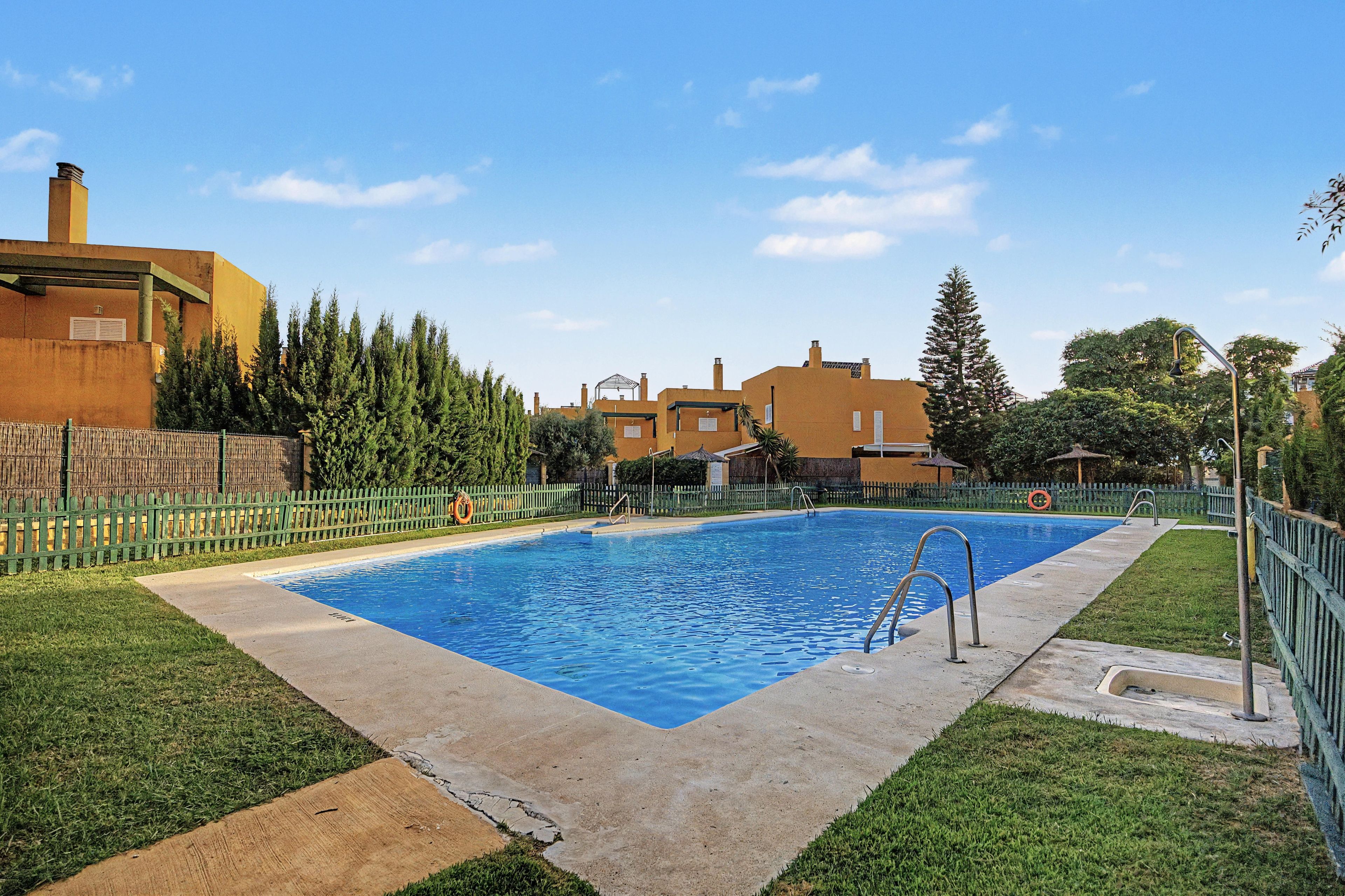 A rectangular swimming pool with a concrete edge, surrounded by a green fence, and a grassy area with a wooden deck.