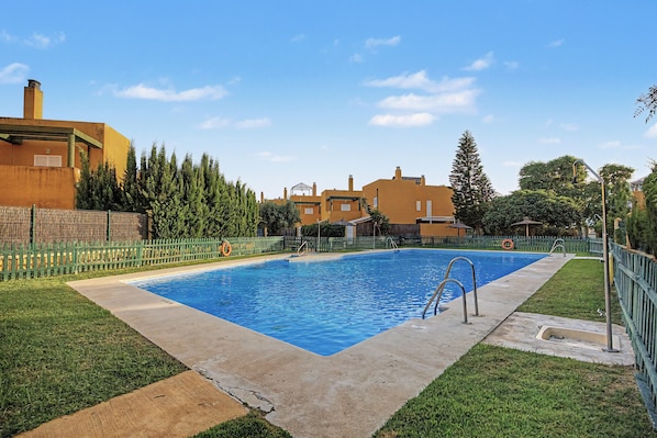 A rectangular swimming pool with a concrete edge, surrounded by a green fence, and a grassy area with a wooden deck. - Casa Tartessos with Pool and Parking in Costa Ballena (Rota)