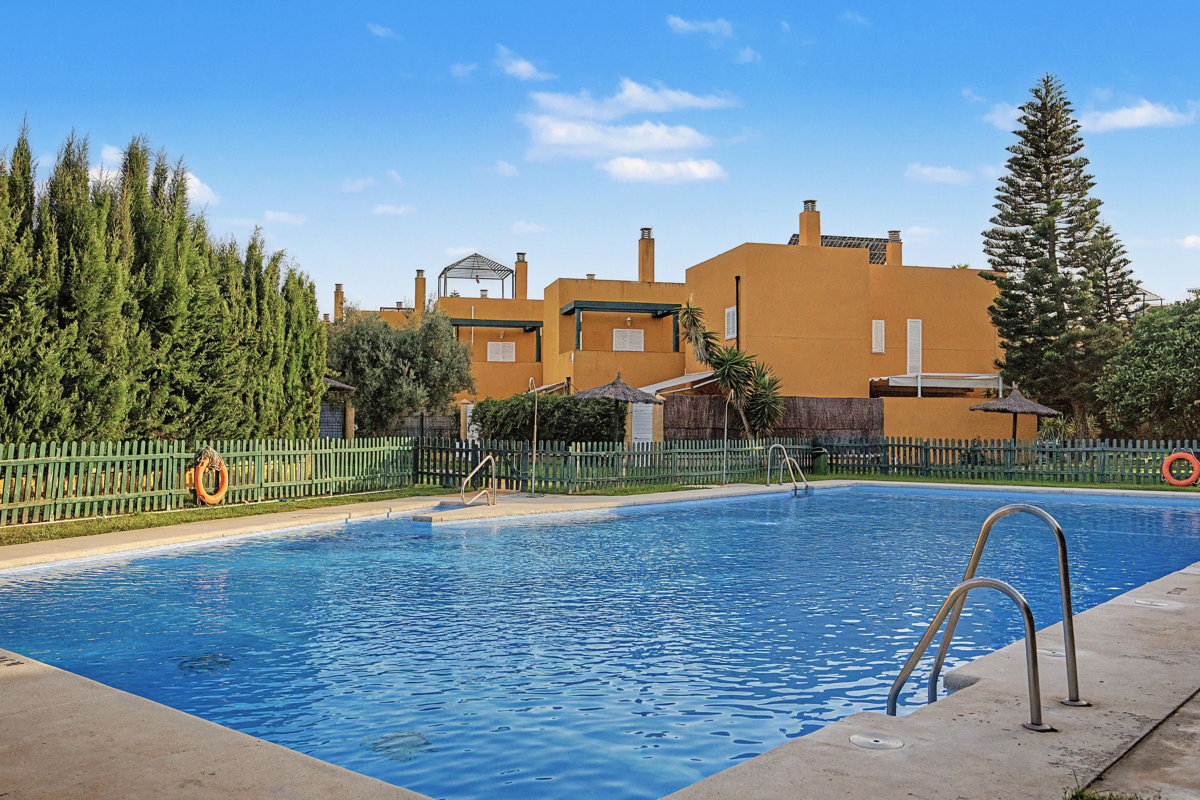 A swimming pool with a metal ladder, surrounded by a fence, and buildings in the background.