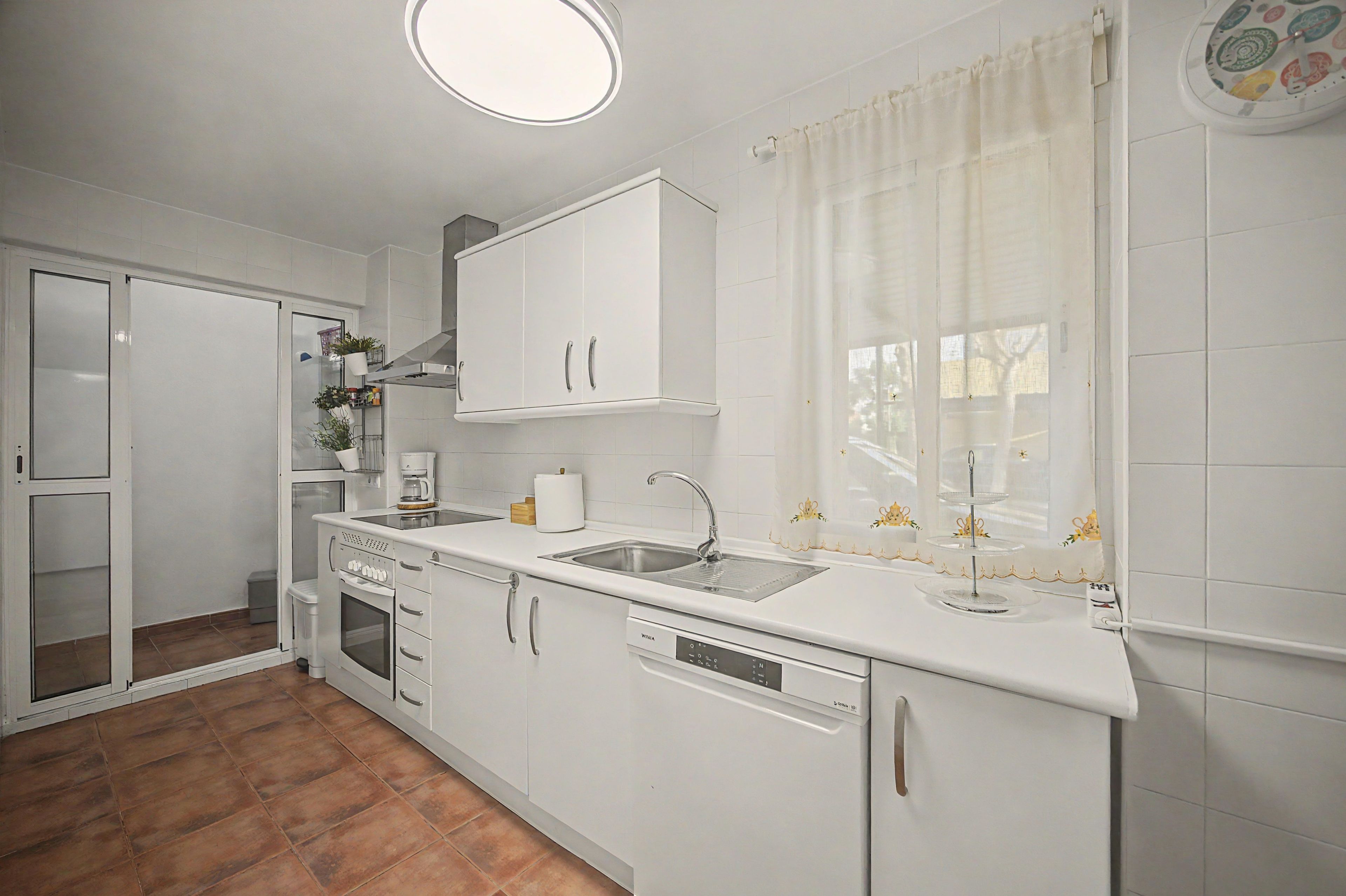 Een moderne keuken met witte kastjes, een inbouwoven, een vaatwasser en een raam met gordijnen.