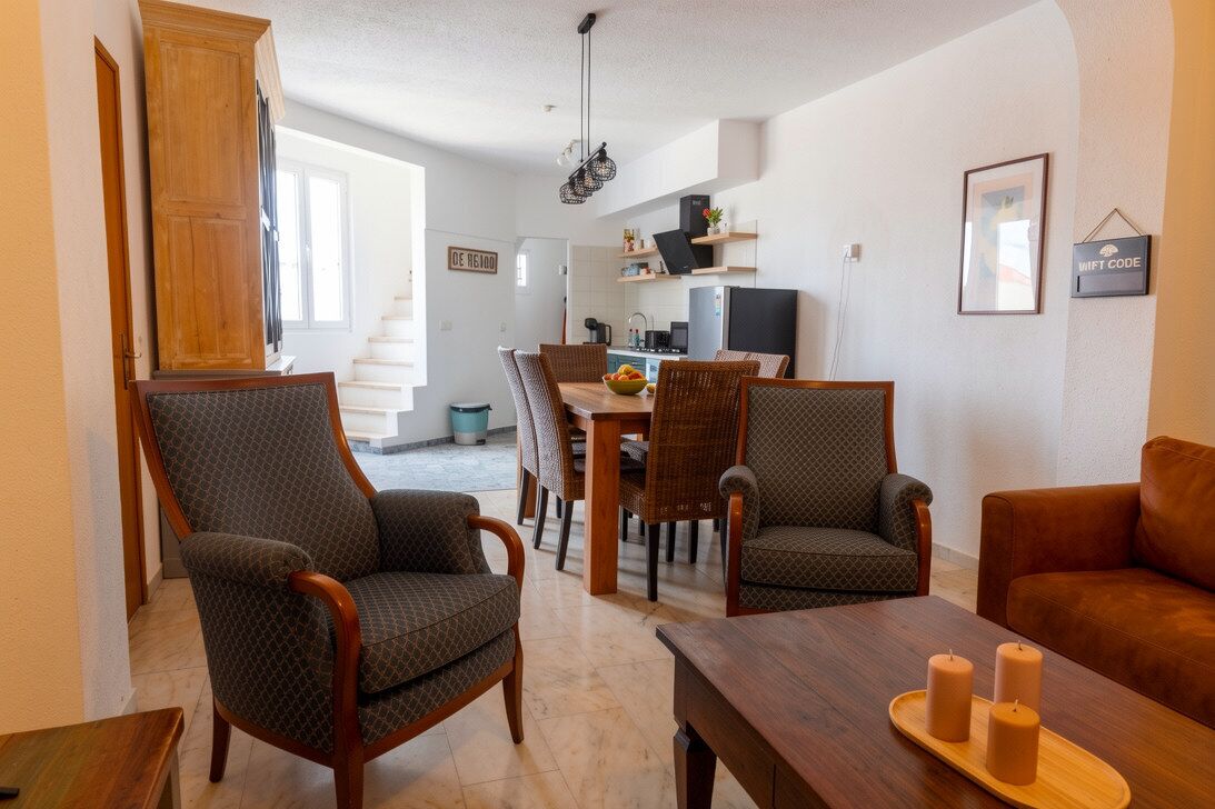 A modern living room with a dining area, featuring a wooden cabinet, wicker chairs, a brown leather sofa, and a wooden coffee table.