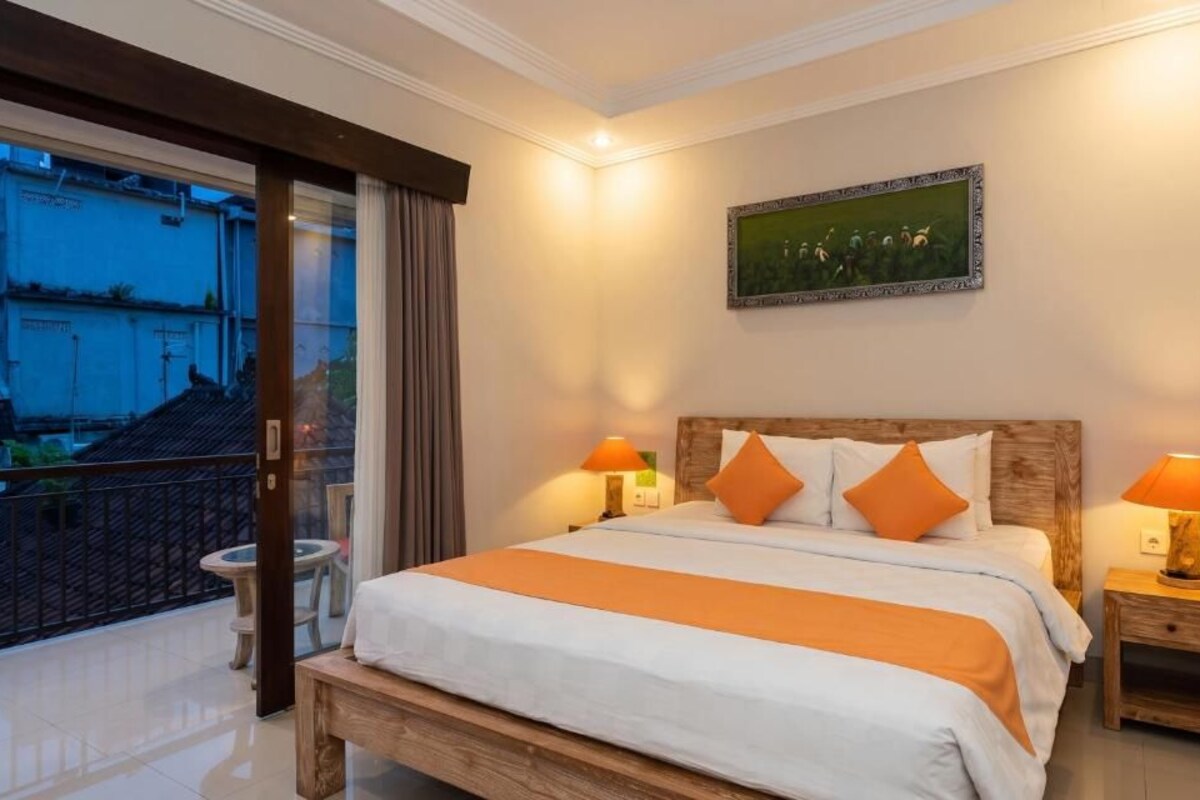 A hotel room with a large bed, two bedside lamps, a painting on the wall, and a balcony with a view of buildings.