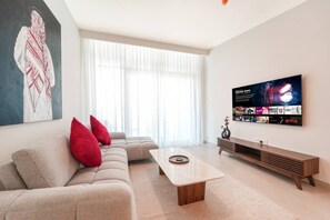 A modern living room with a sofa, red cushions, a coffee table, a TV wall-mounted above a wooden console, and a large window with sheer curtains. - Modern 1BR Retreat in Parkside Views | Dubai Hills (Dubai)