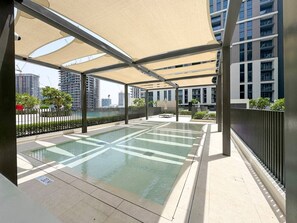 Une piscine extérieure dotée d’un auvent, entourée d’une clôture et bordée de bâtiments modernes en arrière-plan.