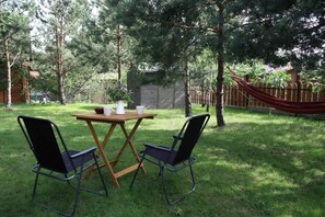 Un espacio al aire libre con mesa de madera y dos sillas, una hamaca y un jardín con césped y árboles.
