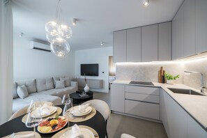 Uma cozinha moderna com eletrodomésticos integrados, um espaço de refeições com mesa redonda posta para uma refeição, e uma sala de estar com sofá.