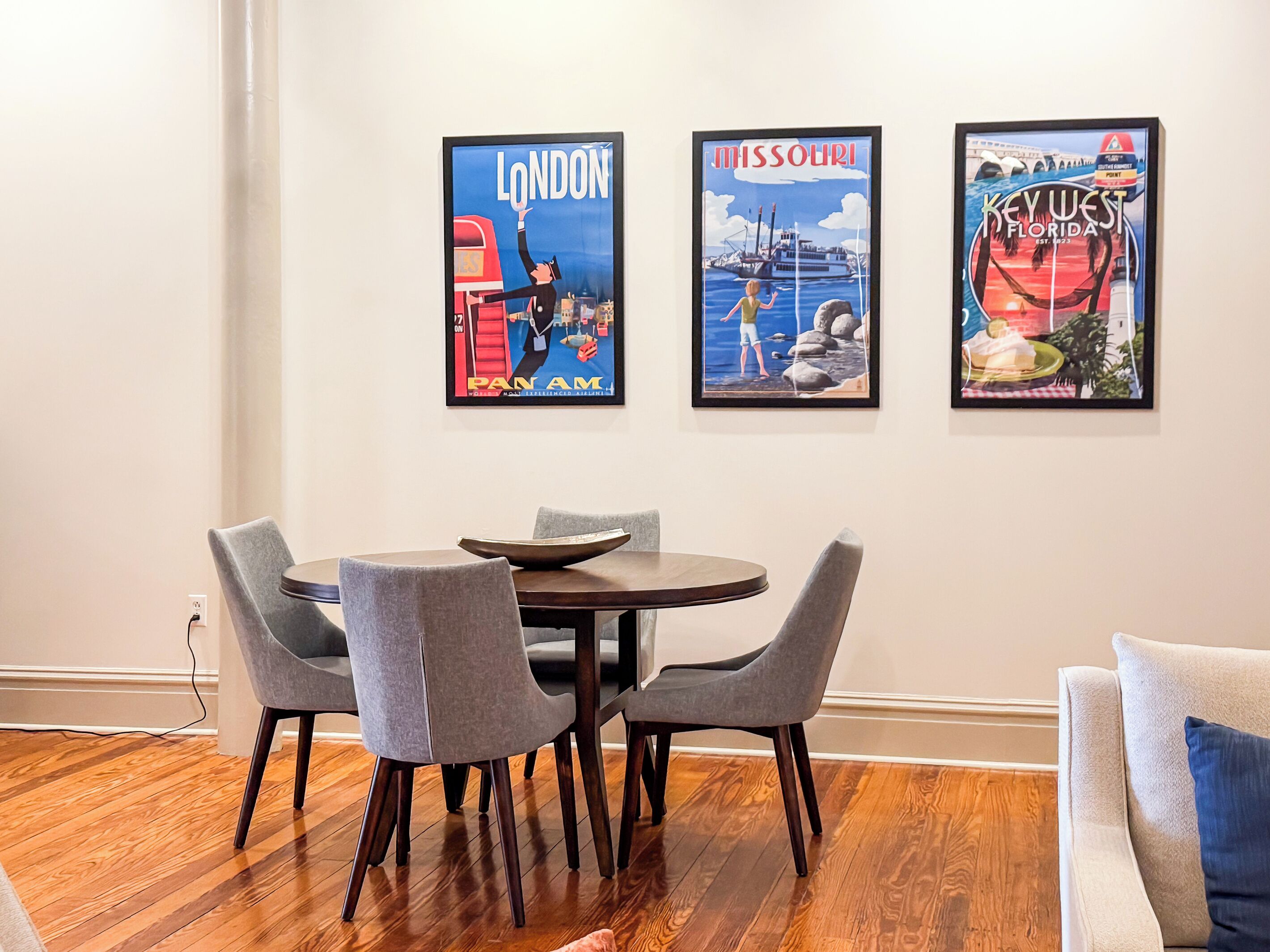 Een eetgedeelte met een ronde tafel en vier stoelen, drie ingelijste reisposters aan de muur en een houten vloer.