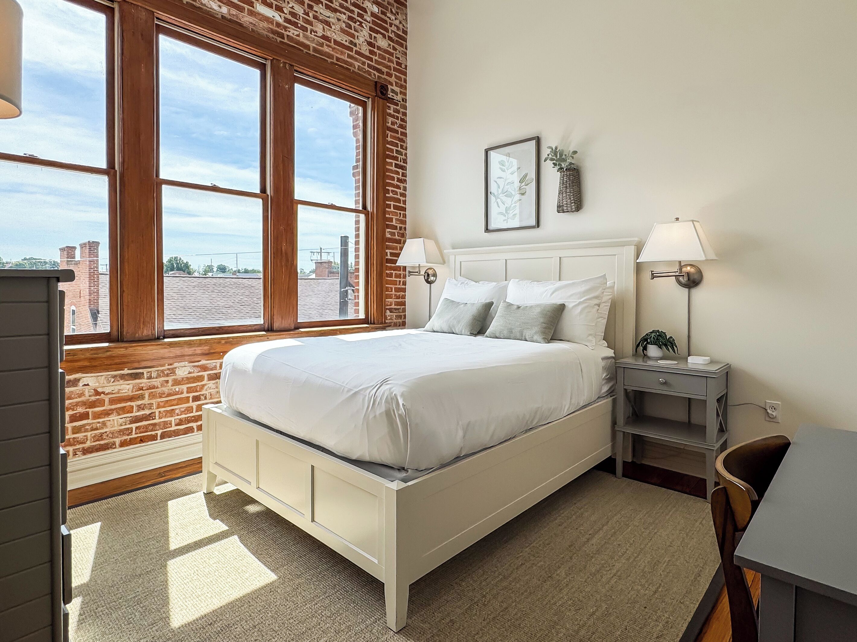 A bedroom with a large window, a bed, a bedside table with a lamp, and a small desk with a chair.