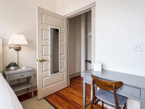 A room with a desk, chair, bedside table with lamp, and a closet.