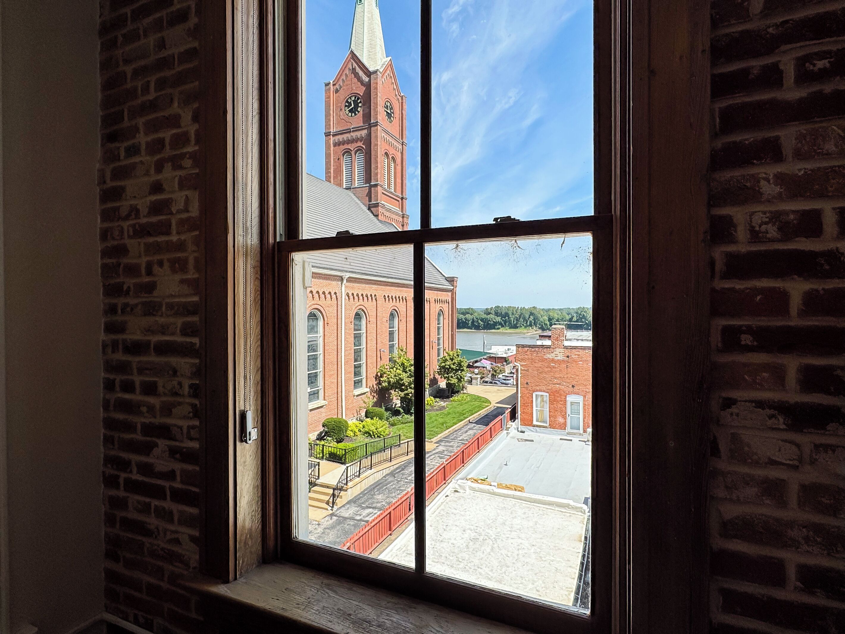 A window with a view of a brick building with a clock tower, a river, and a green park area.