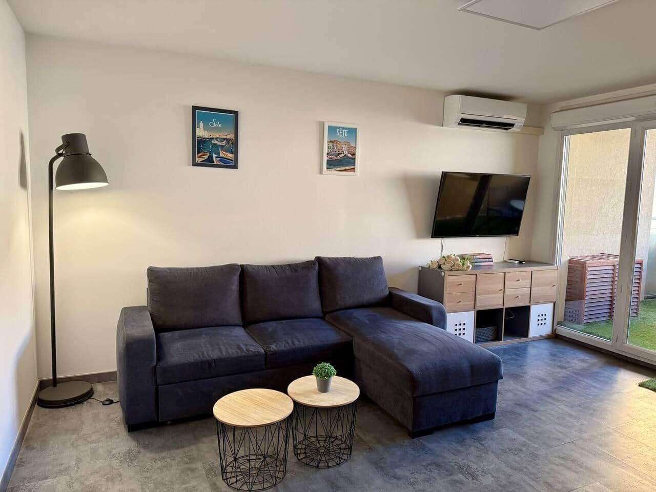 A modern living room with a sectional sofa, two small round tables, a floor lamp, a flat-screen TV, and a sliding glass door leading to an outdoor area.