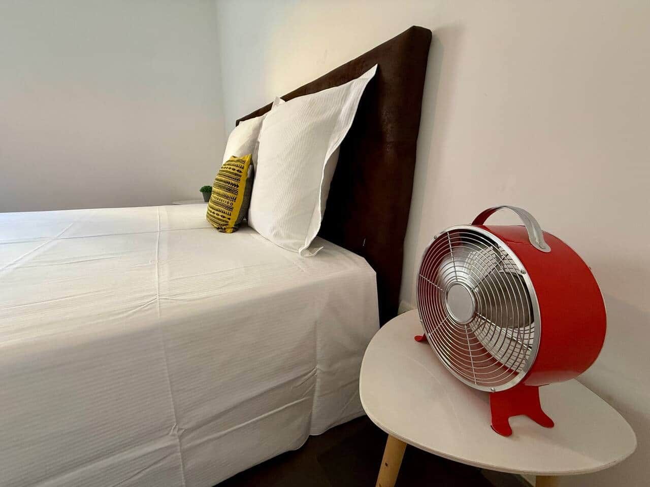 Un lit avec une literie blanche et deux oreillers, un ventilateur rouge posé sur une petite table à côté, et une tête de lit sombre.