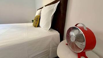 Un lit avec une literie blanche et deux oreillers, un ventilateur rouge posé sur une petite table à côté, et une tête de lit sombre.