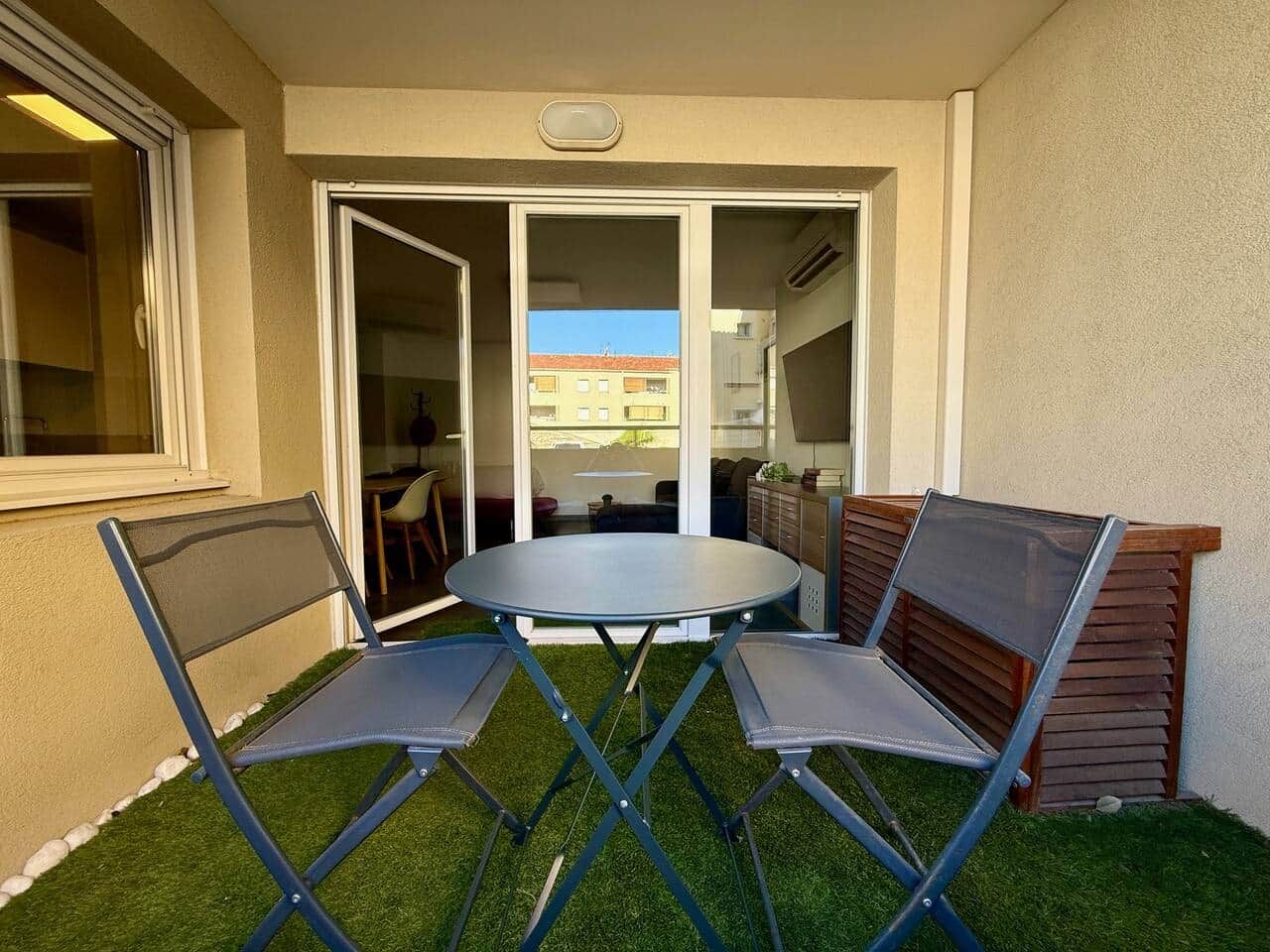 Eine Terrasse mit rundem Tisch und zwei Stühlen, eine Glasschiebetür zu einem Innenbereich und eine kleine Fläche mit Rasenteppich.