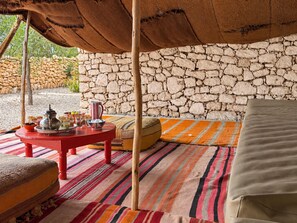Un rincón exterior acogedor con un fondo de pared de piedra, una mesa roja con un juego de té y una alfombra a rayas de colores.