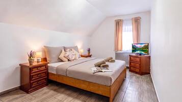 Un dormitorio con una cama de madera, una mesita de noche con lámpara, un aparador, un televisor y un ventanal con cortinas.