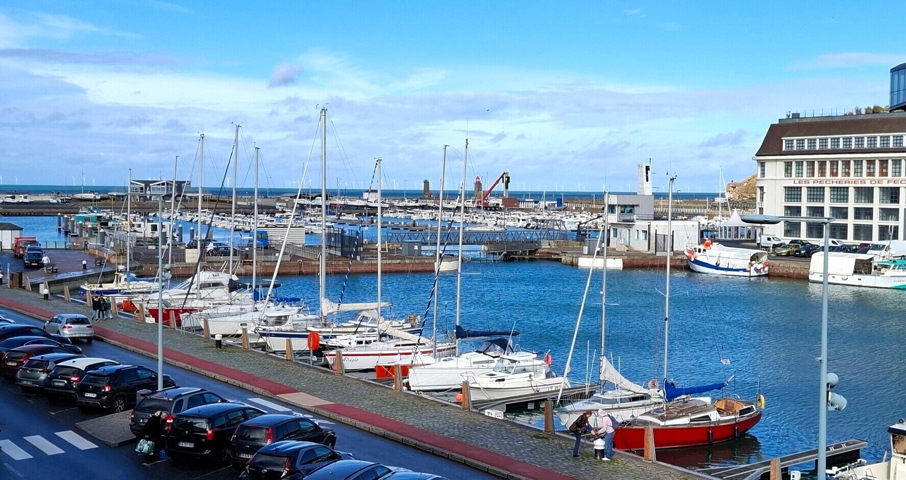 A marina with boats docked, a building labeled 'LES PECHERIES DE FEC,' and a road with cars.