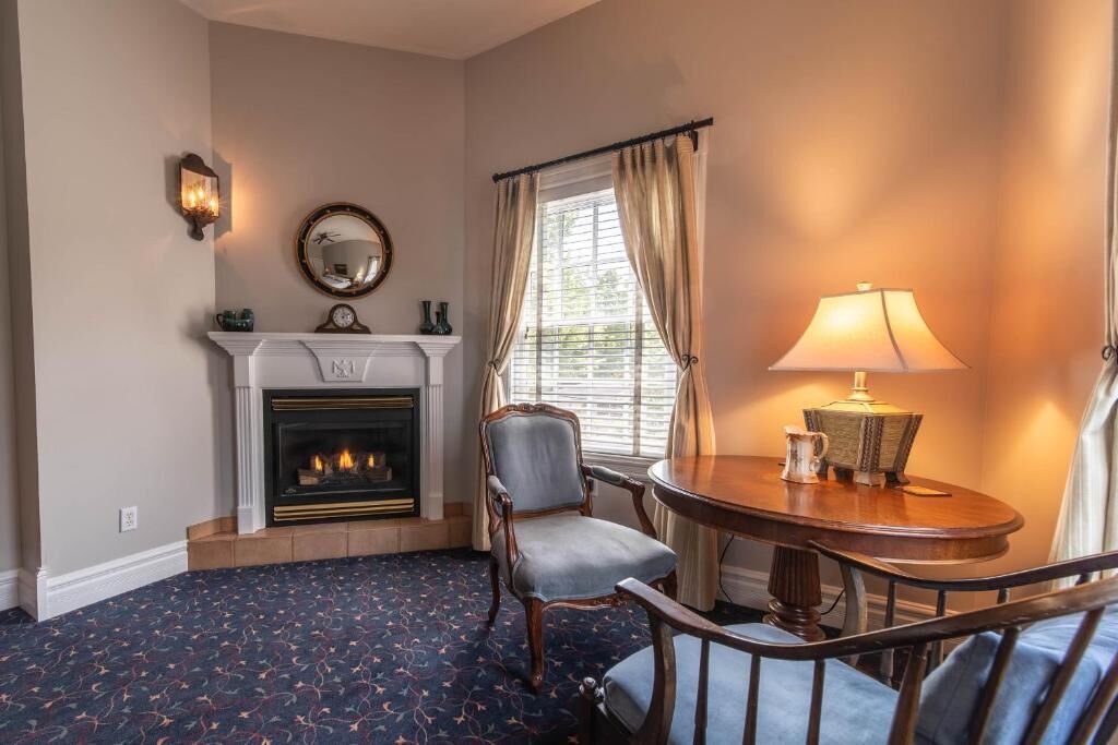 Una habitación con chimenea, una mesa redonda con lámpara, una silla y una ventana con cortinas.