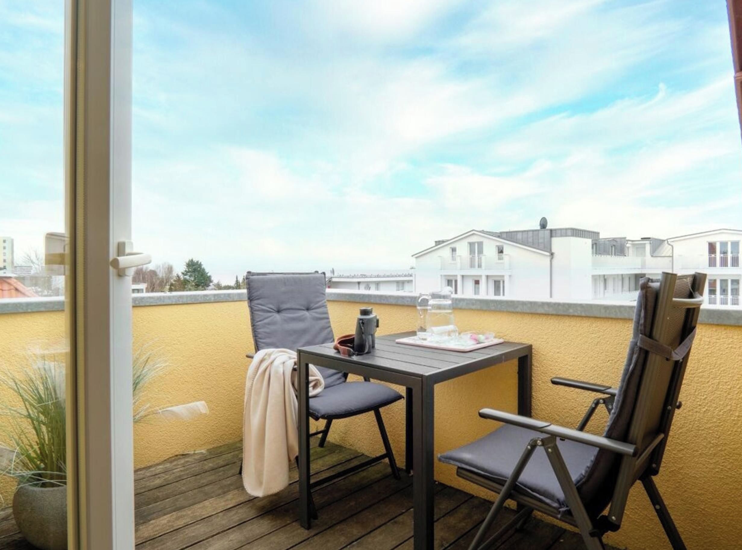 Un balcón con una mesa, dos sillas, una planta en maceta y vistas a edificios.