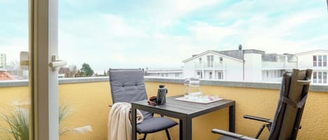 Un balcón con una mesa, dos sillas, una planta en maceta y vistas a edificios.
