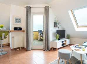 A modern living room with a television, a dining area with a table and chairs, a balcony with a chair, and a large window with curtains. - Holiday Apartment With 1 Bedroom (Grossenbrode)