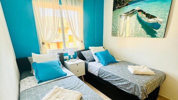 Ein Zimmer mit zwei Betten, einem Deckenventilator, einem Fenster mit Vorhängen und einem Gemälde eines Bootes auf dem Wasser.