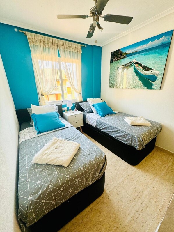 Ein Zimmer mit zwei Betten, einem Deckenventilator, einem Fenster mit Vorhängen und einem Gemälde eines Bootes auf dem Wasser.