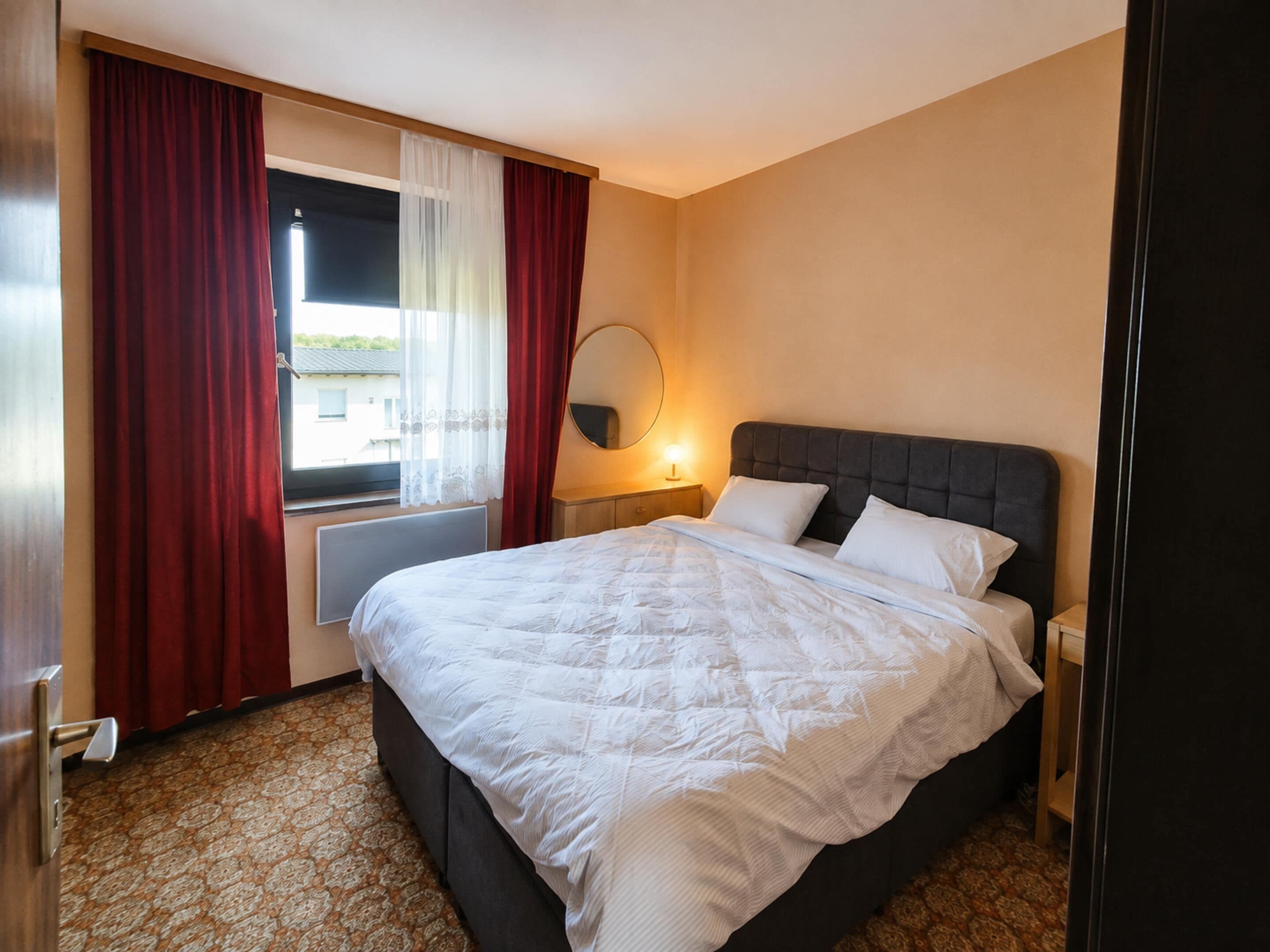 A hotel room with a large bed, a bedside table with a lamp, a window with curtains, and a view of the outside.