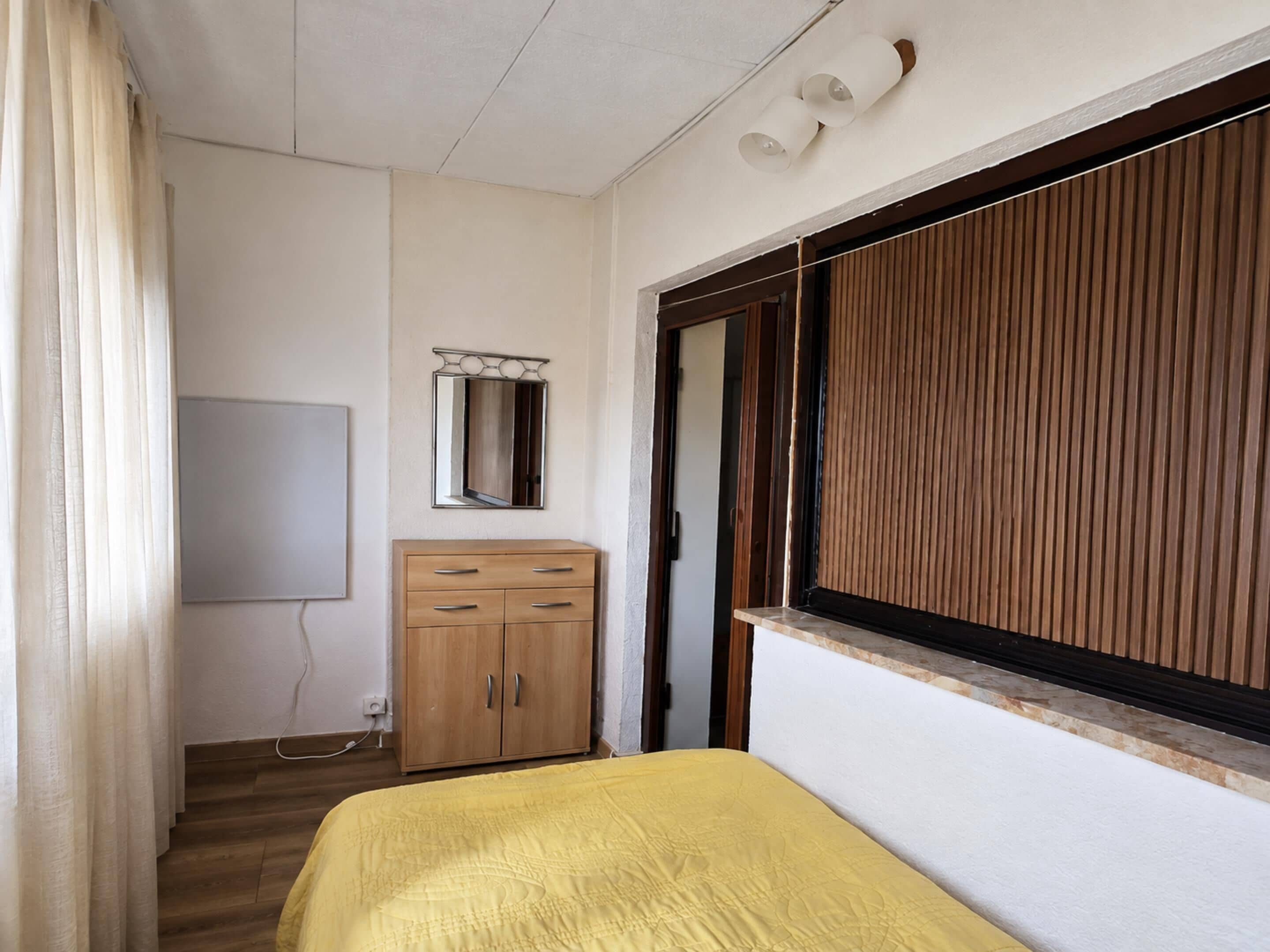Ein Schlafzimmer mit einem Bett, einem Holzschrank, einem Spiegel und einem Fenster mit Jalousien.